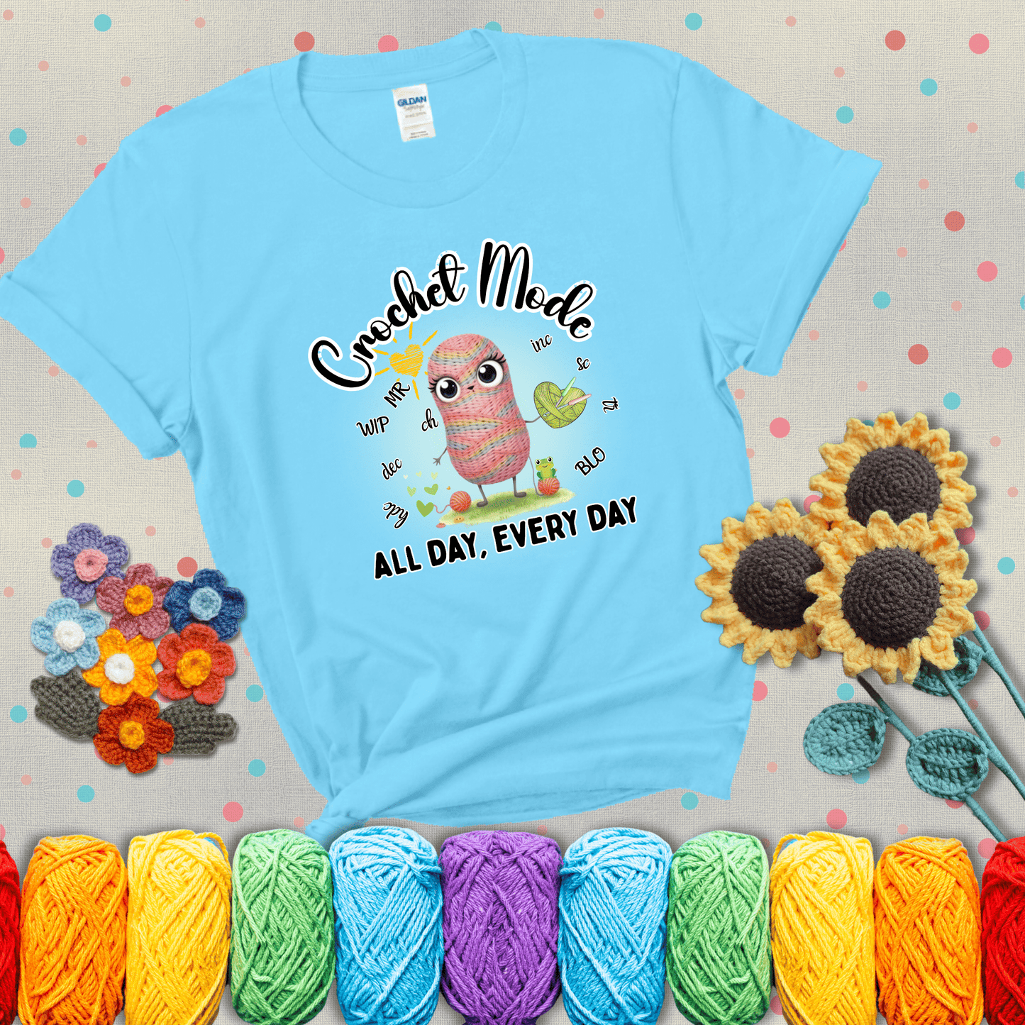 A kawaii-style rainbow yarn character surrounded by crochet stitch abbreviations and holding a yarn heart, with text “Crochet Mode All Day, Every Day"; showing a light blue tee with grey sunflower background