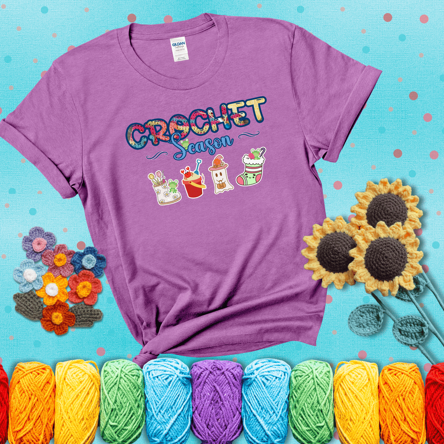 The words “Crochet Season” with cute seasonal icons below: a spring boot with hooks, summer bucket with crab, fall ghost with pumpkin hat, and winter stocking - all with tiny frogs; showing a light purple tee with a teal sunflower background