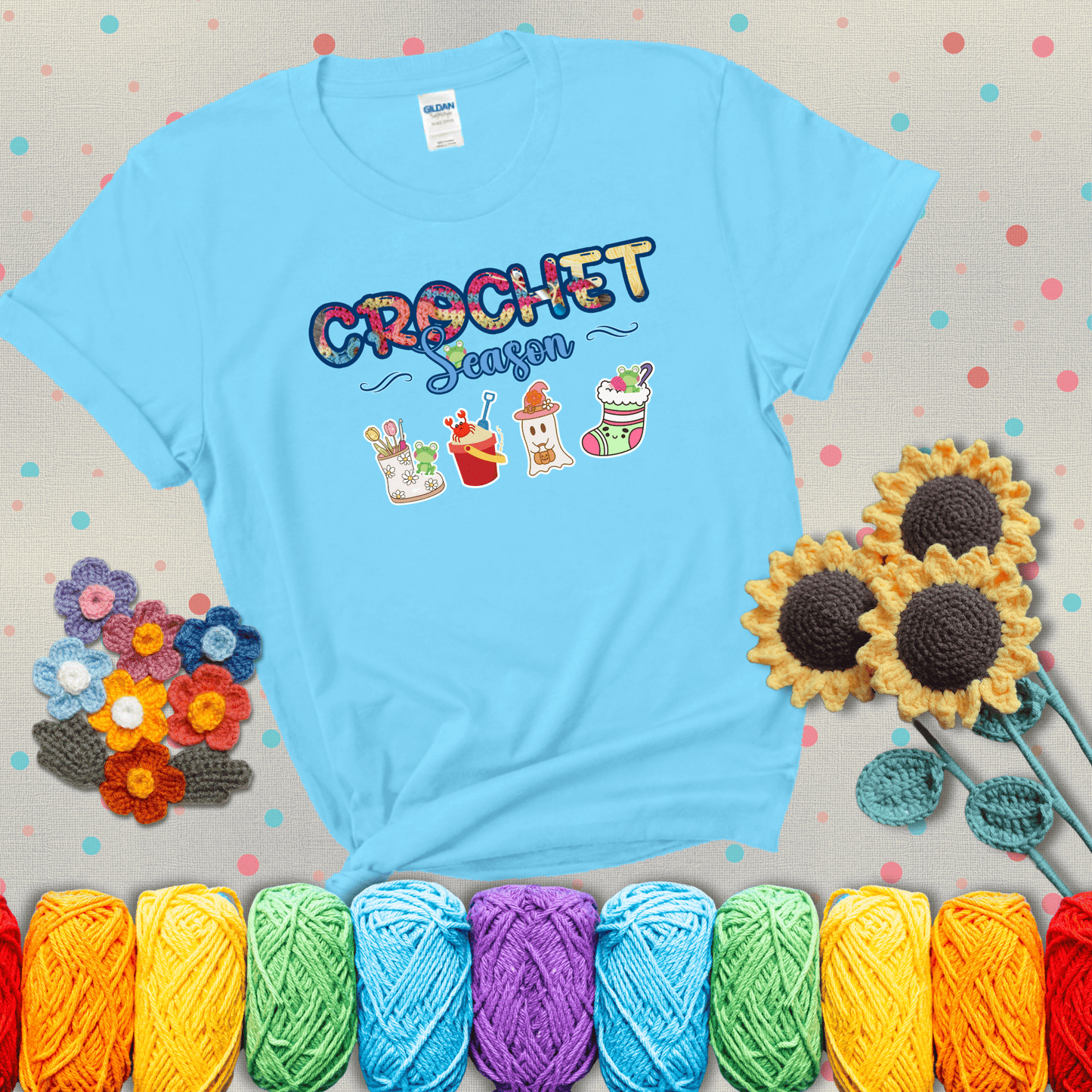 The words “Crochet Season” with cute seasonal icons below: a spring boot with hooks, summer bucket with crab, fall ghost with pumpkin hat, and winter stocking - all with tiny frogs; showing a light blue tee with grey sunflower background