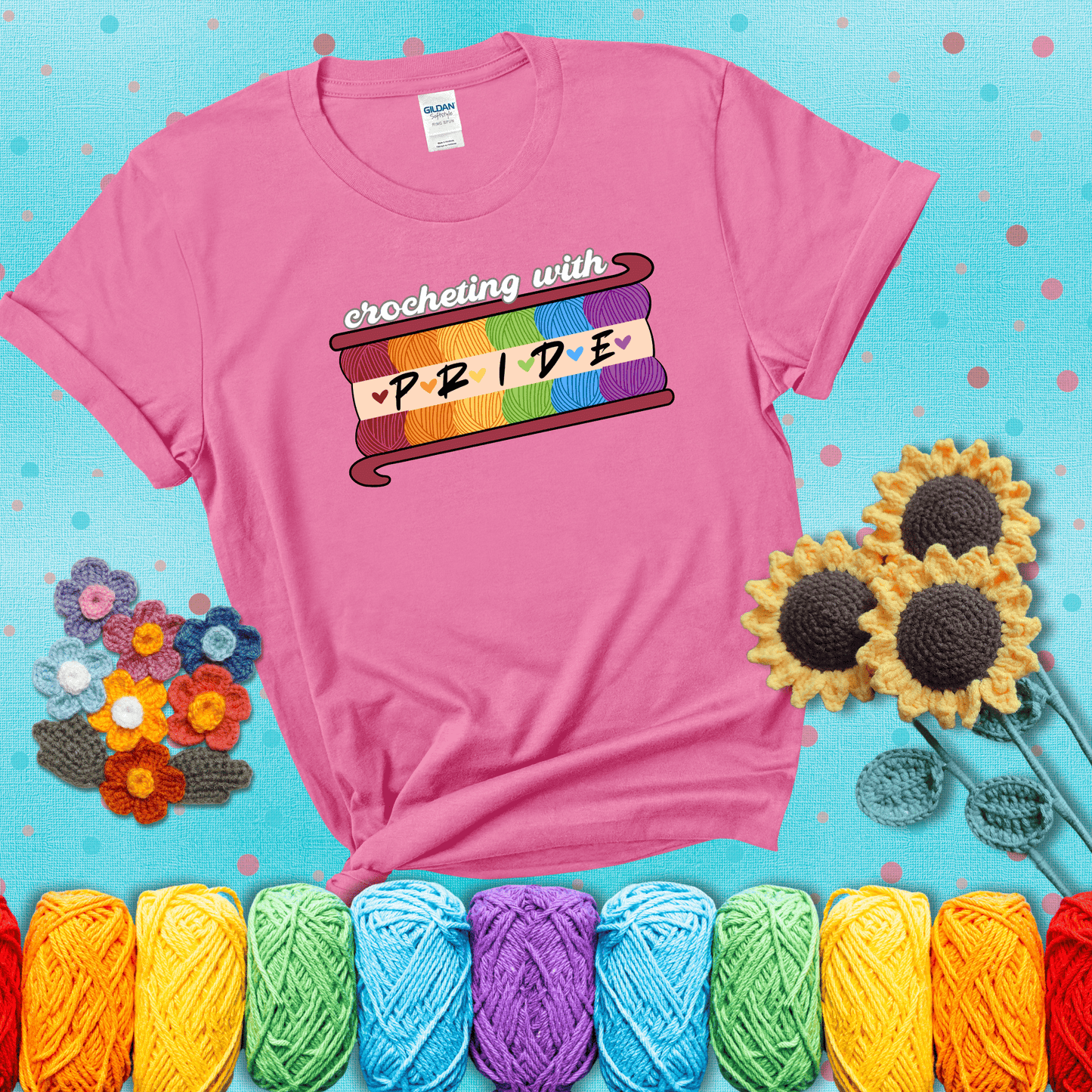 Yarn skeins in rainbow colors stacked in a hook shape. Text reads “Crocheting With Pride” showing a pink tee with a teal sunflower background