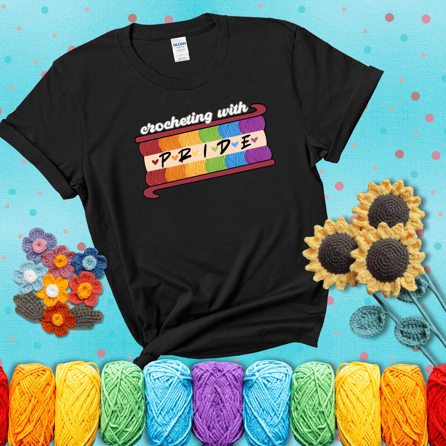 Yarn skeins in rainbow colors stacked in a hook shape. Text reads “Crocheting With Pride” showing a black tee with a teal sunflower background