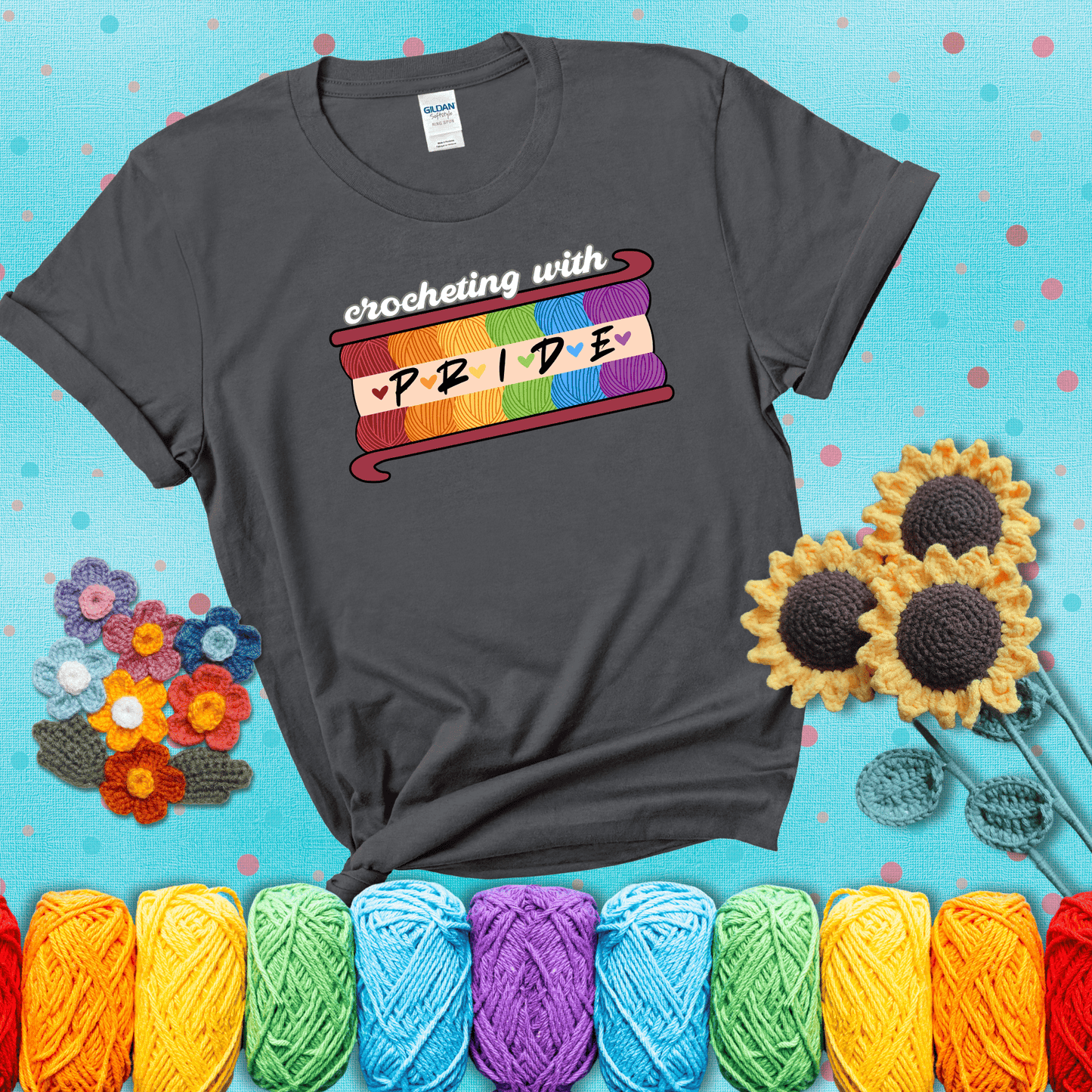 Yarn skeins in rainbow colors stacked in a hook shape. Text reads “Crocheting With Pride” showing a dark grey tee with a teal sunflower background