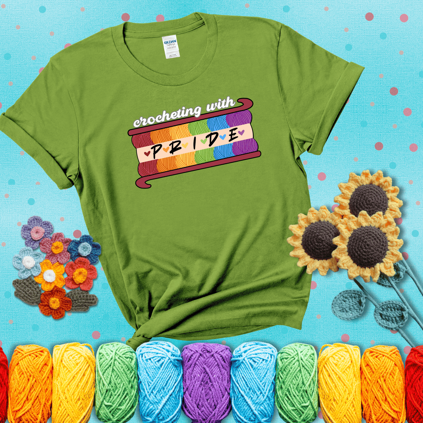 Yarn skeins in rainbow colors stacked in a hook shape. Text reads “Crocheting With Pride” showing a bright green tee with a teal sunflower background