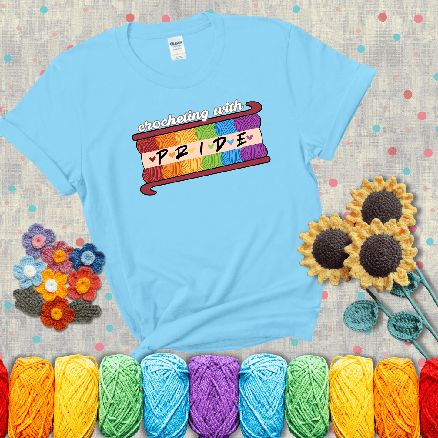 Yarn skeins in rainbow colors stacked in a hook shape. Text reads “Crocheting With Pride” showing a light blue tee with grey sunflower background