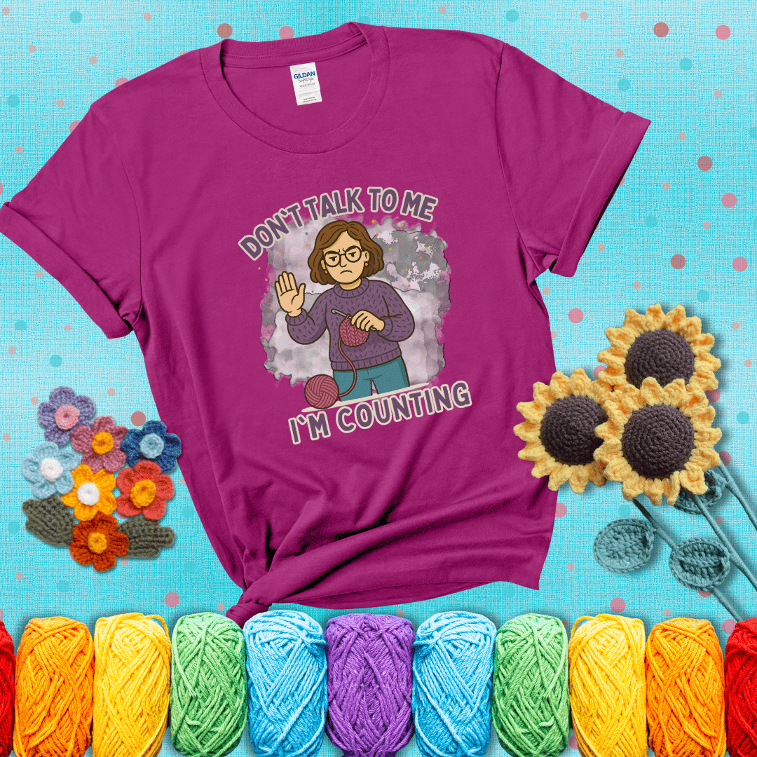 Annoyed expression with yarn and a hand held up to the viewer. Text reads: “Don’t Talk to Me, I’m Counting.” A relatable tee for focused crocheters and knitters alike, showing a magenta tee with a teal sunflower background