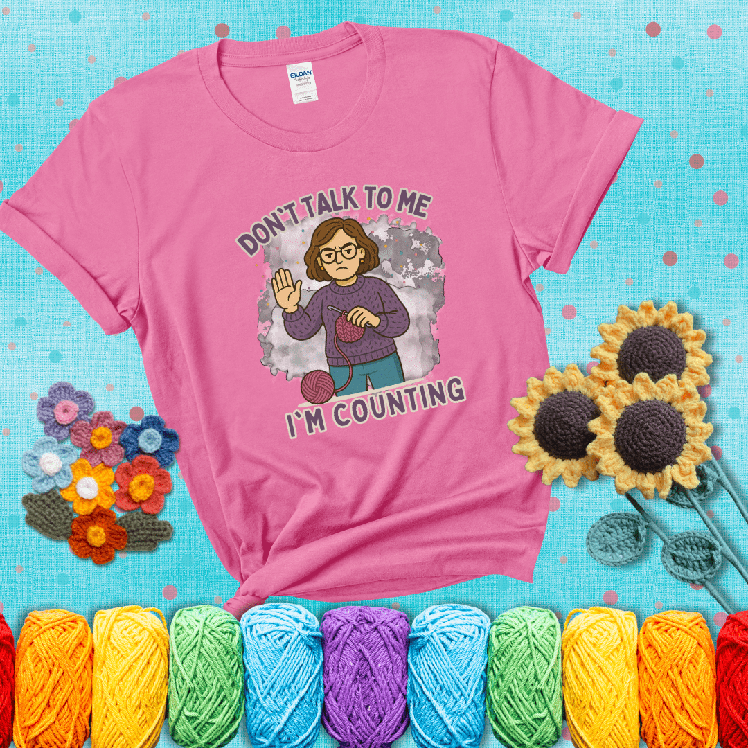 Annoyed expression with yarn and a hand held up to the viewer. Text reads: “Don’t Talk to Me, I’m Counting.” A relatable tee for focused crocheters and knitters alike, showing a pink tee with a teal sunflower background