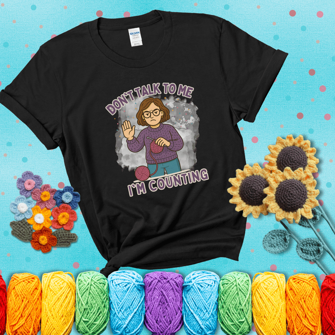 Annoyed expression with yarn and a hand held up to the viewer. Text reads: “Don’t Talk to Me, I’m Counting.” A relatable tee for focused crocheters and knitters alike, showing a black tee with a teal sunflower background