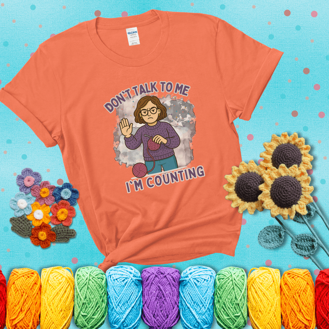 Annoyed expression with yarn and a hand held up to the viewer. Text reads: “Don’t Talk to Me, I’m Counting.” A relatable tee for focused crocheters and knitters alike, showing a light orange tee with a teal sunflower background