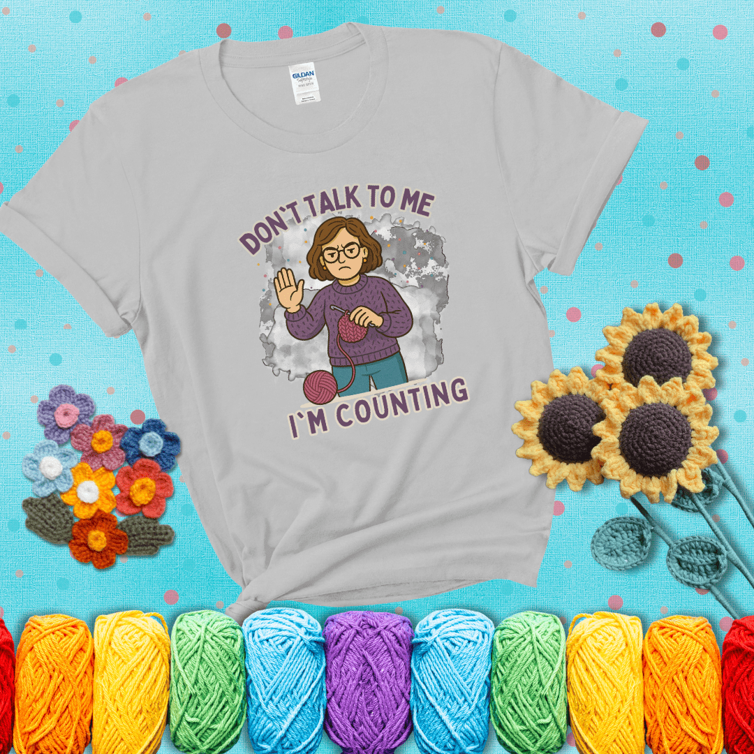 Annoyed expression with yarn and a hand held up to the viewer. Text reads: “Don’t Talk to Me, I’m Counting.” A relatable tee for focused crocheters and knitters alike, showing a light grey tee with a teal sunflower background