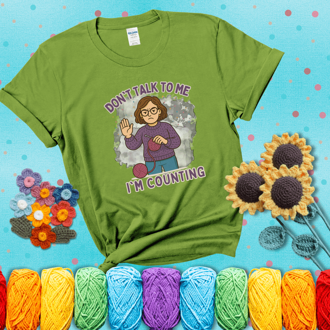 Annoyed expression with yarn and a hand held up to the viewer. Text reads: “Don’t Talk to Me, I’m Counting.” A relatable tee for focused crocheters and knitters alike, showing a bright green tee with a teal sunflower background