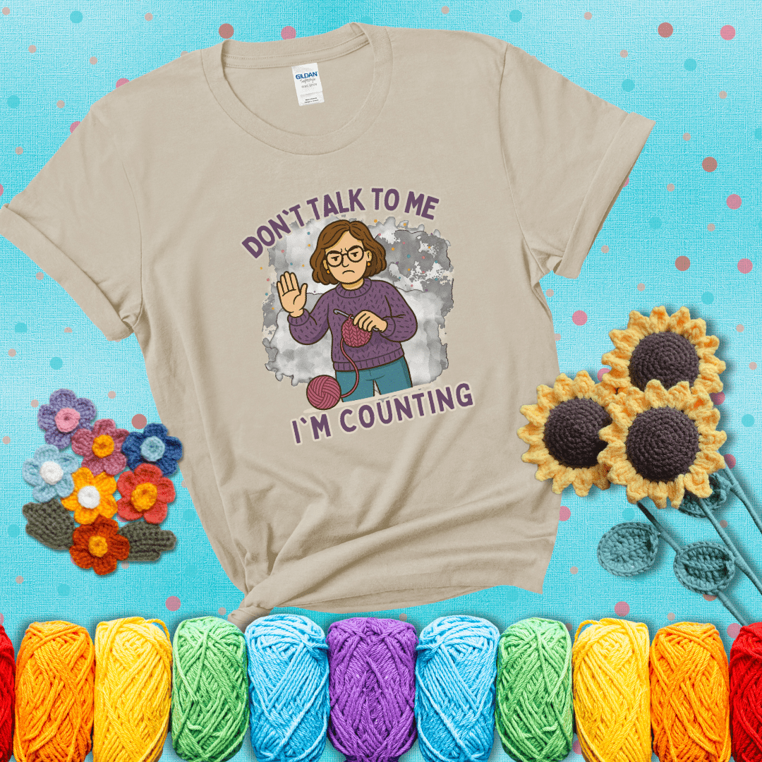 Annoyed expression with yarn and a hand held up to the viewer. Text reads: “Don’t Talk to Me, I’m Counting.” A relatable tee for focused crocheters and knitters alike, showing a beige tee with a teal sunflower background