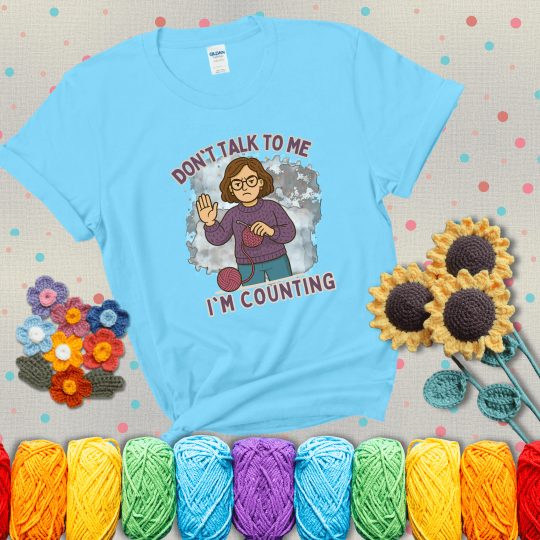 Annoyed expression with yarn and a hand held up to the viewer. Text reads: “Don’t Talk to Me, I’m Counting.” A relatable tee for focused crocheters and knitters alike, showing a light blue tee with grey sunflower background