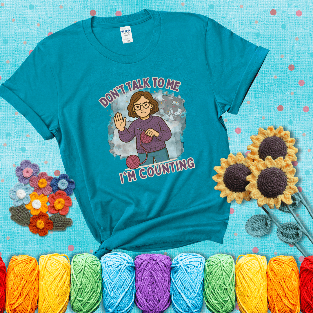 Annoyed expression with yarn and a hand held up to the viewer. Text reads: “Don’t Talk to Me, I’m Counting.” A relatable tee for focused crocheters and knitters alike, showing a blue-green tee with a teal sunflower background