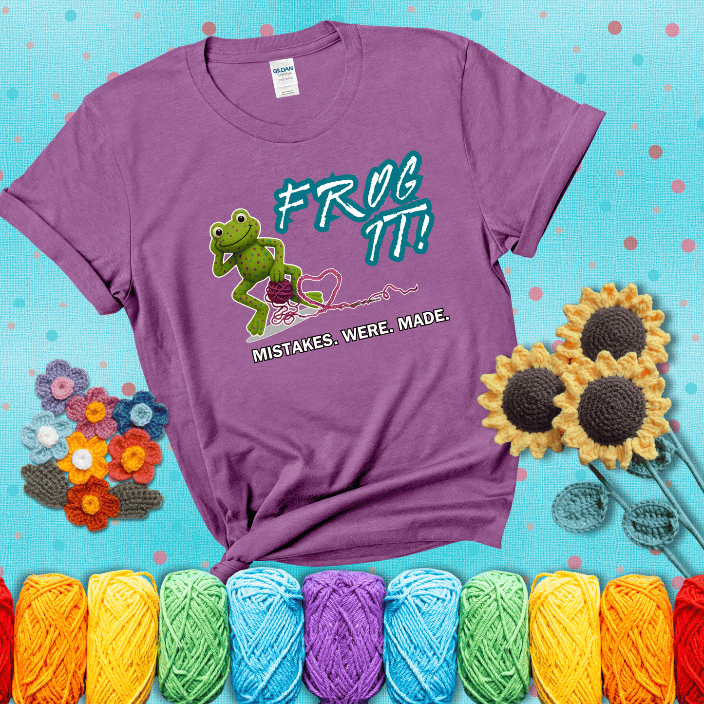 Green frog with yarn and heart-shaped loop. Text reads: “Frog It! Mistakes Were Made.” A playful crochet tee for anyone who's ever had to rip it back, showing a light purple tee with a teal sunflower background