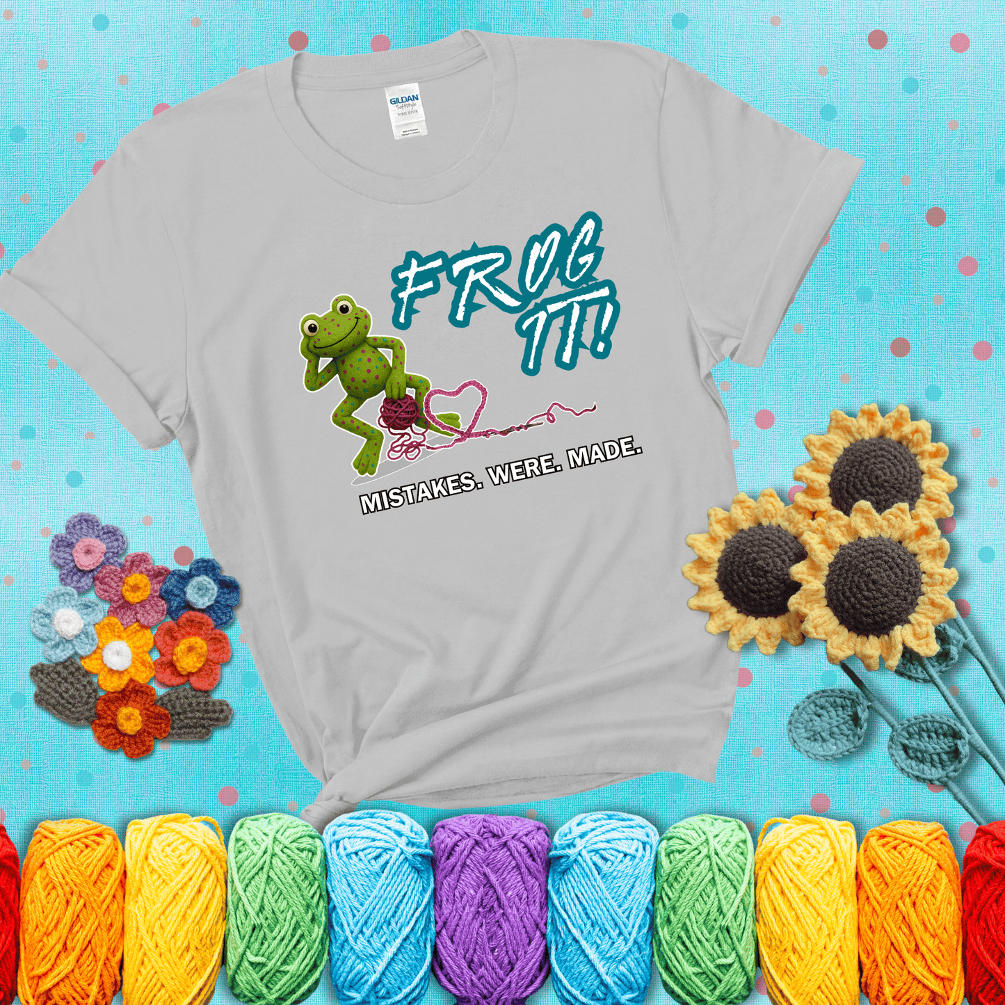 Green frog with yarn and heart-shaped loop. Text reads: “Frog It! Mistakes Were Made.” A playful crochet tee for anyone who's ever had to rip it back, showing a light grey tee with a teal sunflower background