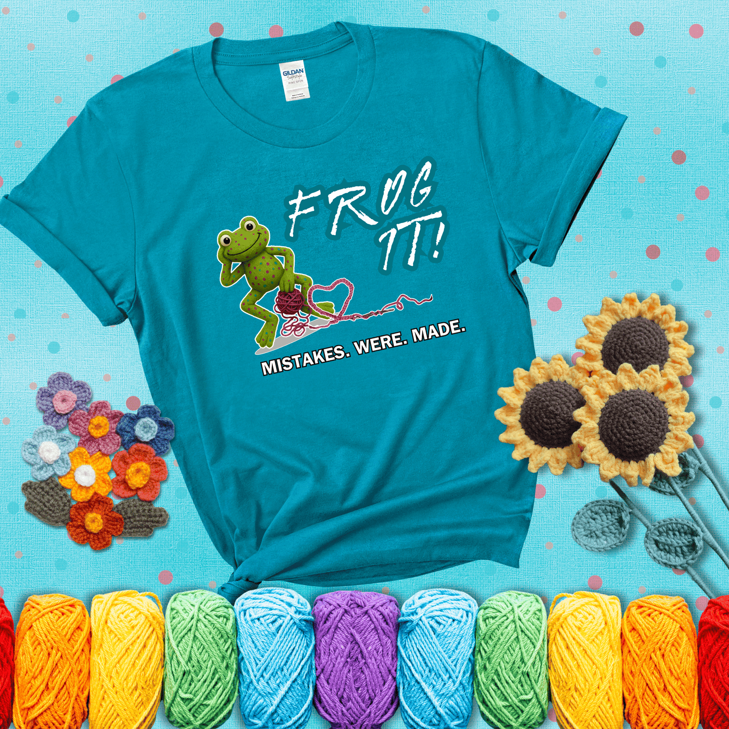 Green frog with yarn and heart-shaped loop. Text reads: “Frog It! Mistakes Were Made.” A playful crochet tee for anyone who's ever had to rip it back, showing a blue-green tee with a teal sunflower background
