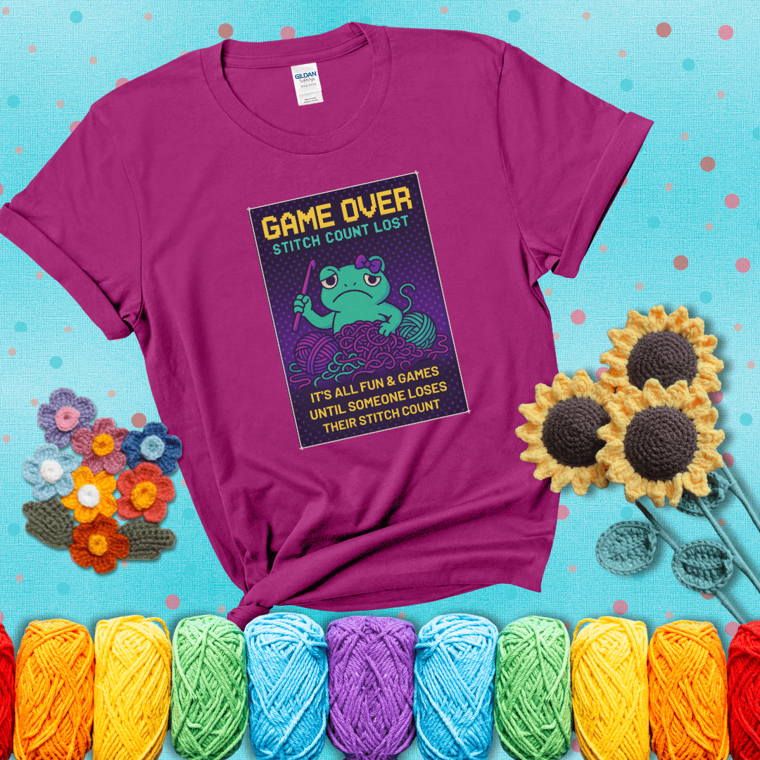 Pixel-style design of a grumpy yarn frog holding a hook, surrounded by yarn chaos, with the text “Game Over. Stitch Count Lost” showing a magenta tee with a teal sunflower background