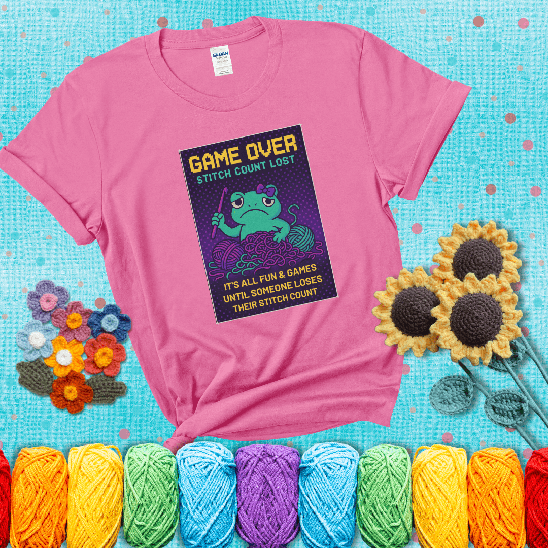 Pixel-style design of a grumpy yarn frog holding a hook, surrounded by yarn chaos, with the text “Game Over. Stitch Count Lost” showing a pink tee with a teal sunflower background