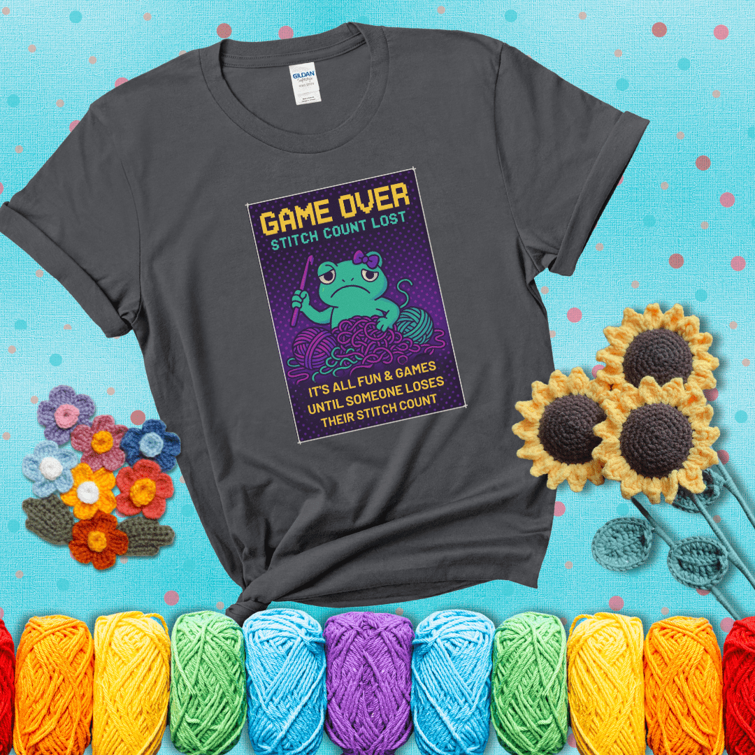 Pixel-style design of a grumpy yarn frog holding a hook, surrounded by yarn chaos, with the text “Game Over. Stitch Count Lost” showing a dark grey tee with a teal sunflower background