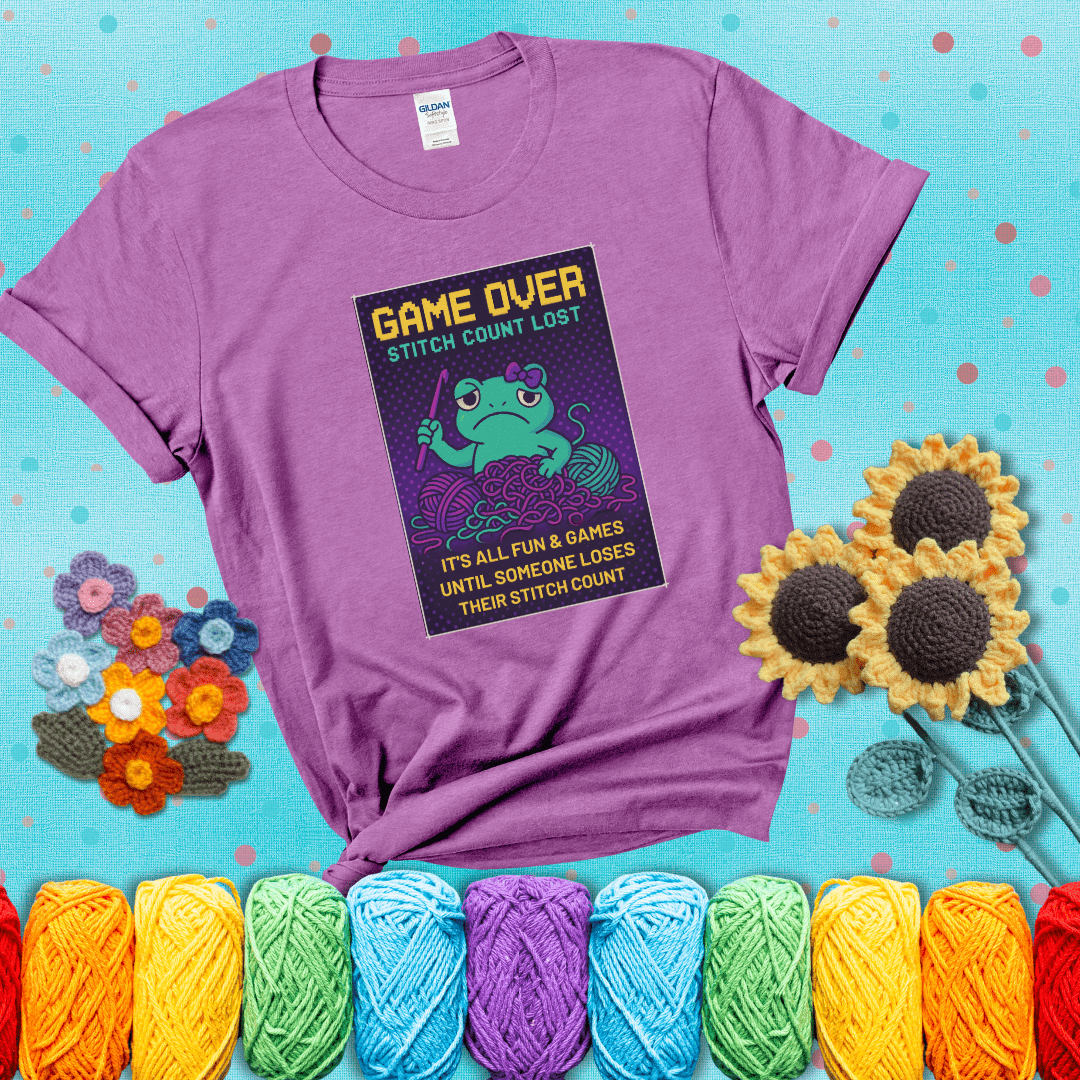 Pixel-style design of a grumpy yarn frog holding a hook, surrounded by yarn chaos, with the text “Game Over. Stitch Count Lost” showing a light purple tee with a teal sunflower background