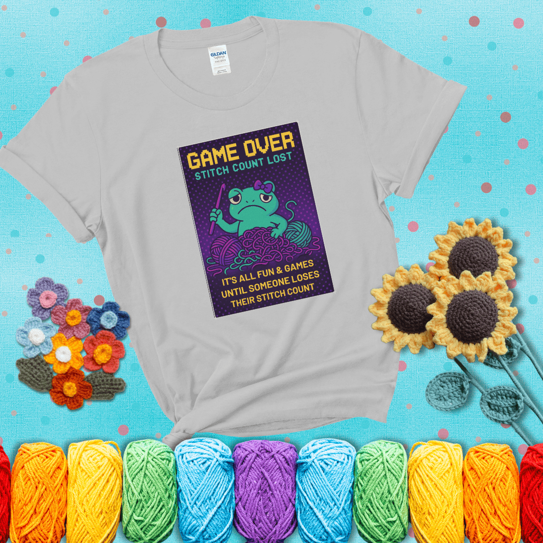 Pixel-style design of a grumpy yarn frog holding a hook, surrounded by yarn chaos, with the text “Game Over. Stitch Count Lost” showing a light grey tee with a teal sunflower background