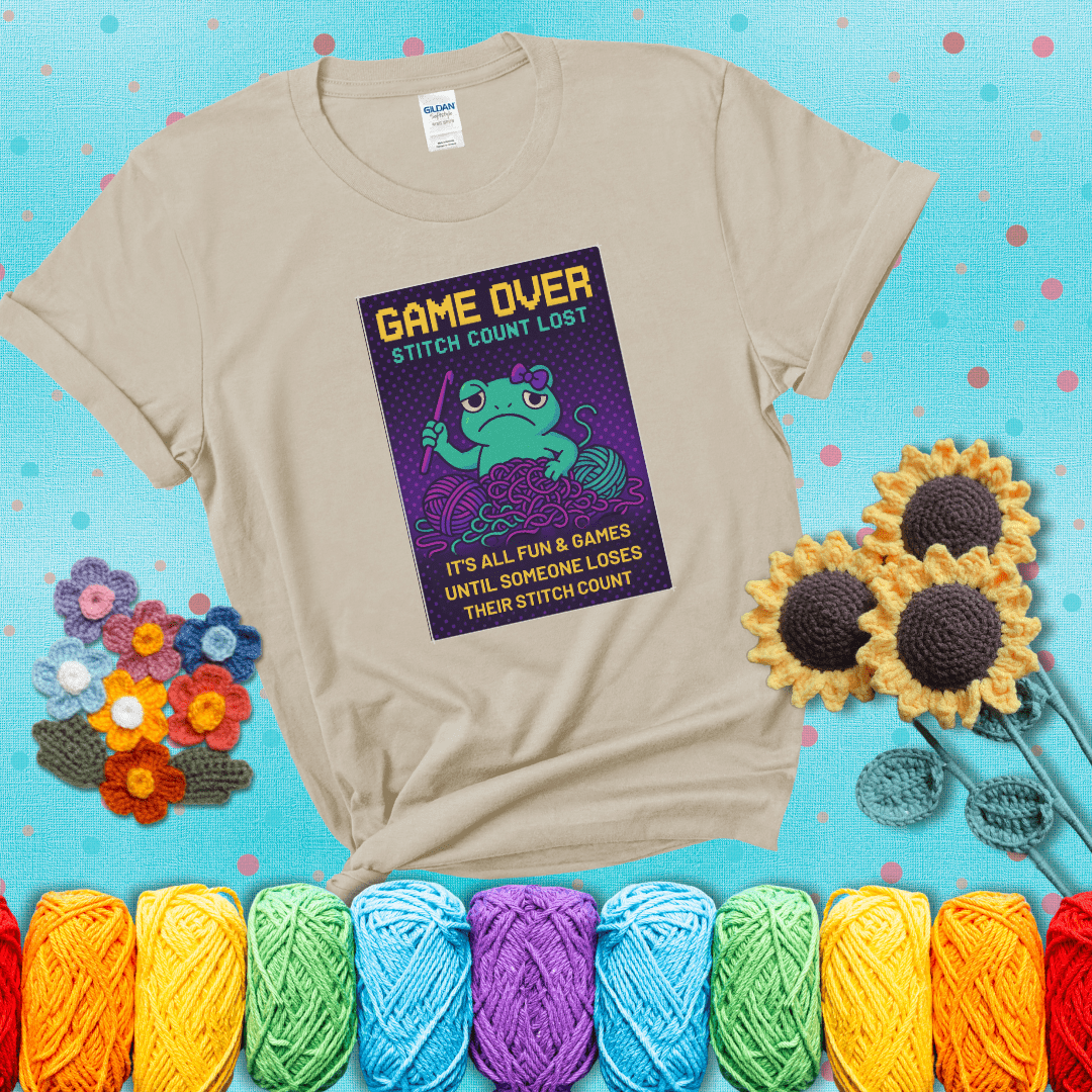 Pixel-style design of a grumpy yarn frog holding a hook, surrounded by yarn chaos, with the text “Game Over. Stitch Count Lost” showing a beige tee with a teal sunflower background