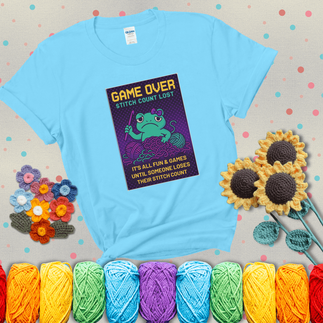 Pixel-style design of a grumpy yarn frog holding a hook, surrounded by yarn chaos, with the text “Game Over. Stitch Count Lost” showing a light blue tee with grey sunflower background