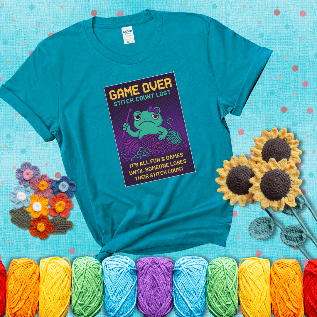 Pixel-style design of a grumpy yarn frog holding a hook, surrounded by yarn chaos, with the text “Game Over. Stitch Count Lost” showing a blue-green tee with a teal sunflower background