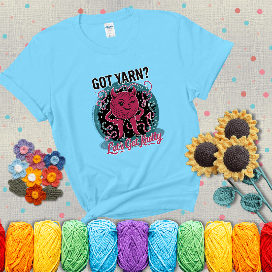 A cheeky pink yarn ball with devil horns, arms, and legs winks under the text “Got Yarn? Let’s Get Knotty,” showing a light blue tee with grey sunflower background