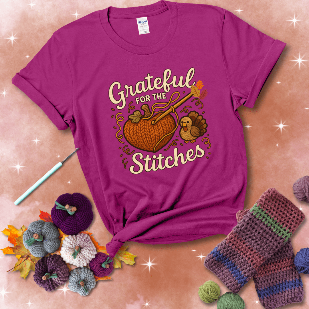 A cozy fall scene with a crochet hook stitching a heart-shaped pumpkin, surrounded by yarn and a small turkey, showing a magenta tee with a festive Fall background