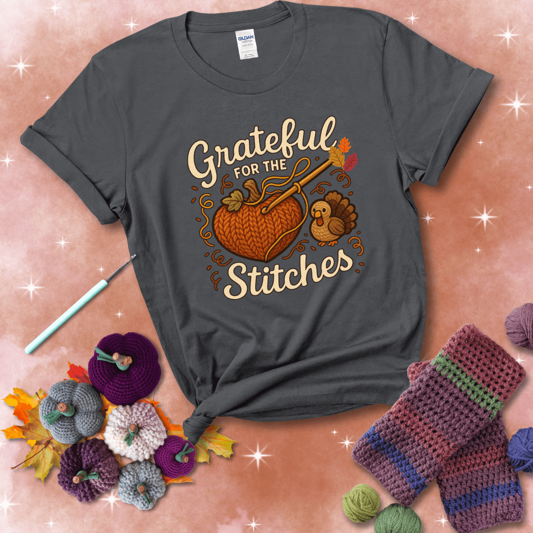 A cozy fall scene with a crochet hook stitching a heart-shaped pumpkin, surrounded by yarn and a small turkey, showing a dark grey tee with a festive Fall background