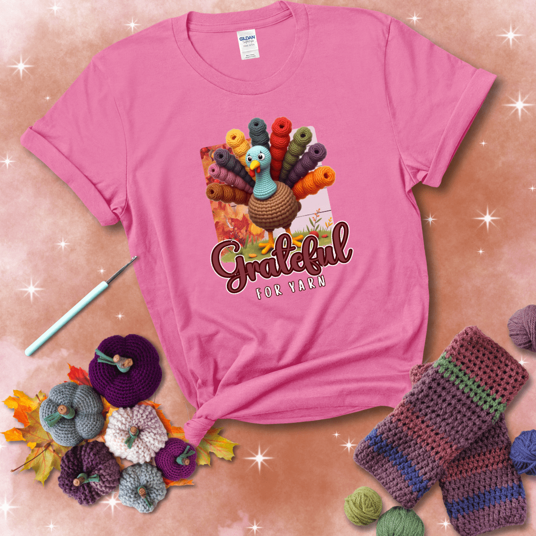 A crocheted turkey with colorful yarn skeins for tail feathers, standing on fall leaves with the text “Grateful for Yarn” showing a pink tee with a festive Fall background