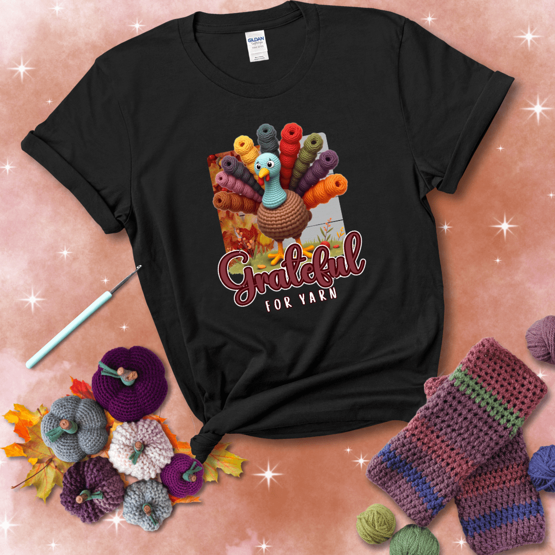 A crocheted turkey with colorful yarn skeins for tail feathers, standing on fall leaves with the text “Grateful for Yarn” showing a black tee with a festive Fall background