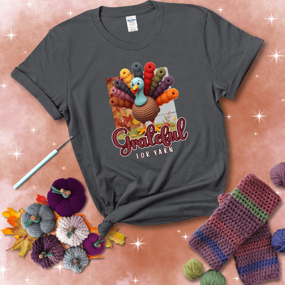 A crocheted turkey with colorful yarn skeins for tail feathers, standing on fall leaves with the text “Grateful for Yarn” showing a dark grey tee with a festive Fall background