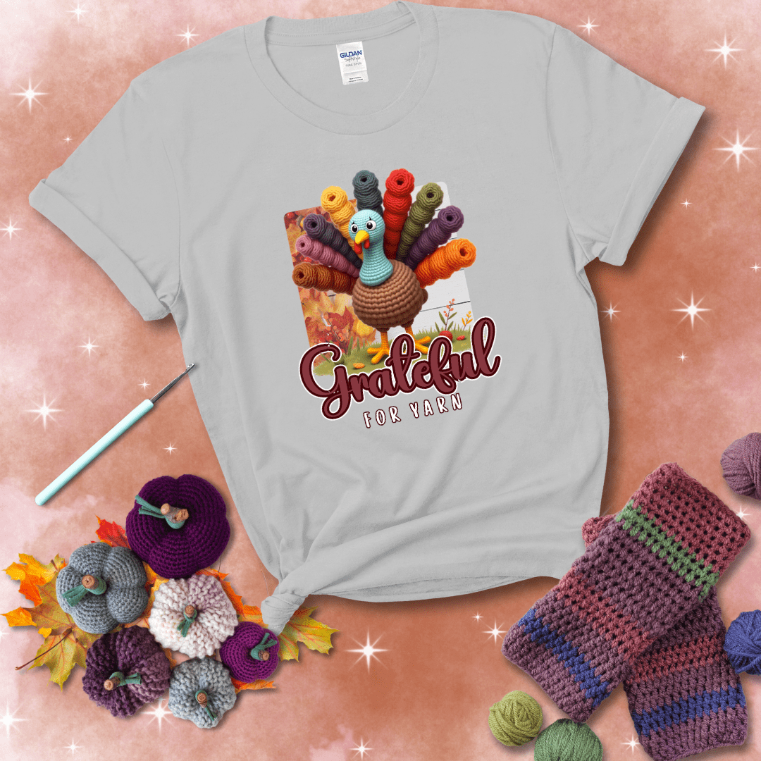 A crocheted turkey with colorful yarn skeins for tail feathers, standing on fall leaves with the text “Grateful for Yarn” showing a light grey tee with a festive Fall background
