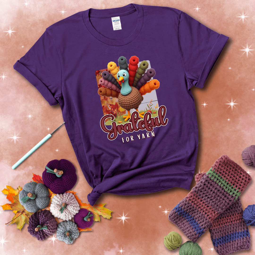 A crocheted turkey with colorful yarn skeins for tail feathers, standing on fall leaves with the text “Grateful for Yarn” showing a purple tee with a festive Fall background