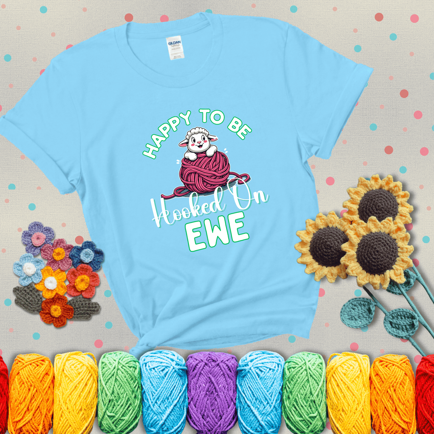 Smiling cartoon sheep peeks from a pink yarn ball, surrounded by green text “Happy to Be Hooked on Ewe” — playful and affectionate sheep pun for crocheters showing a light blue tee with grey sunflower background