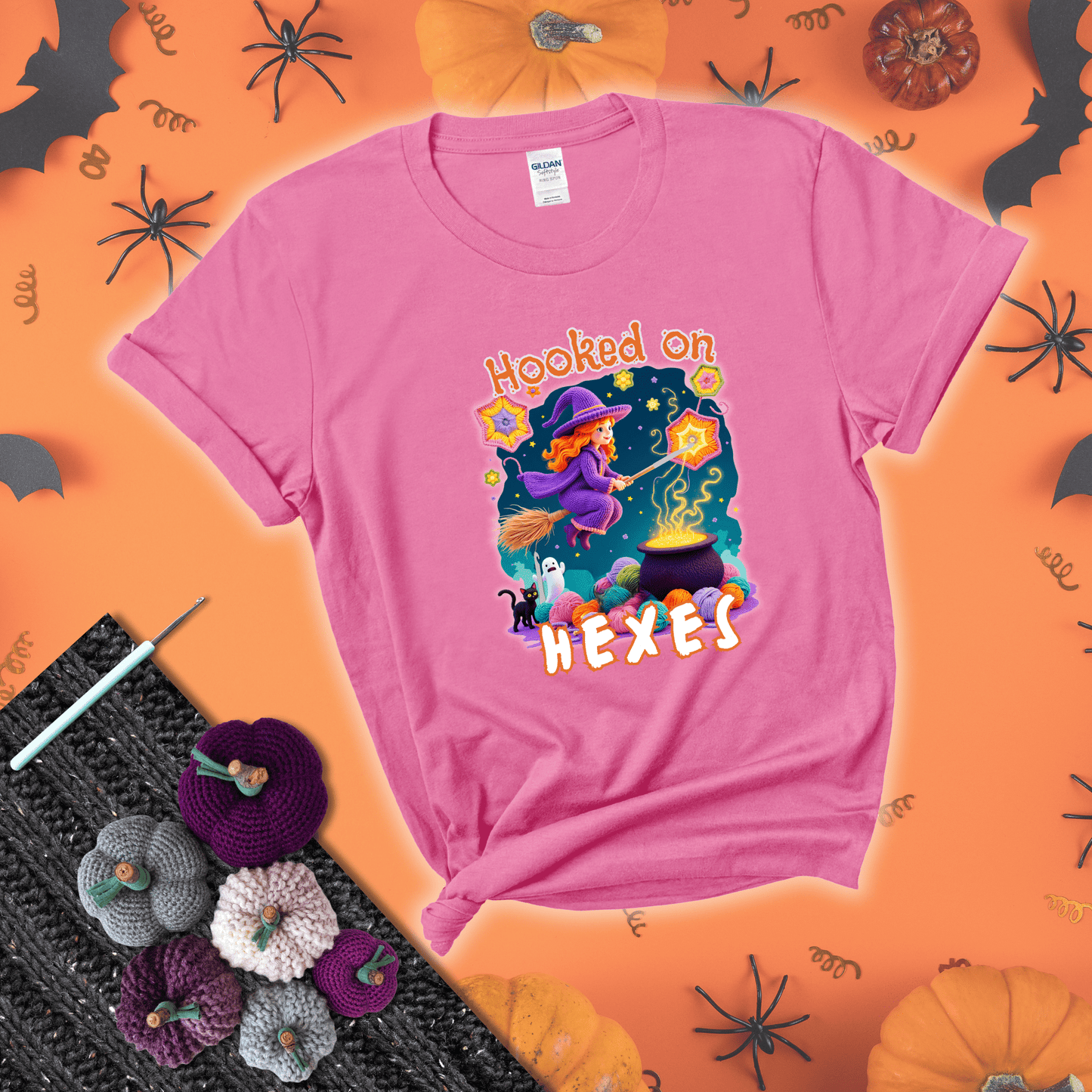 Witchy vibe with stars, hexagons, and a crochet hook. Text reads: “Hooked on Hexes.” A mystical and moody tee for fans of spooky stitches showing a pink tee with a festive Halloween background