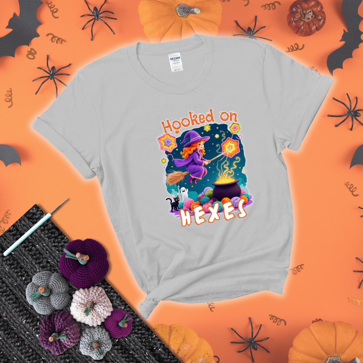 Witchy vibe with stars, hexagons, and a crochet hook. Text reads: “Hooked on Hexes.” A mystical and moody tee for fans of spooky stitches showing a light grey tee with a festive Halloween background