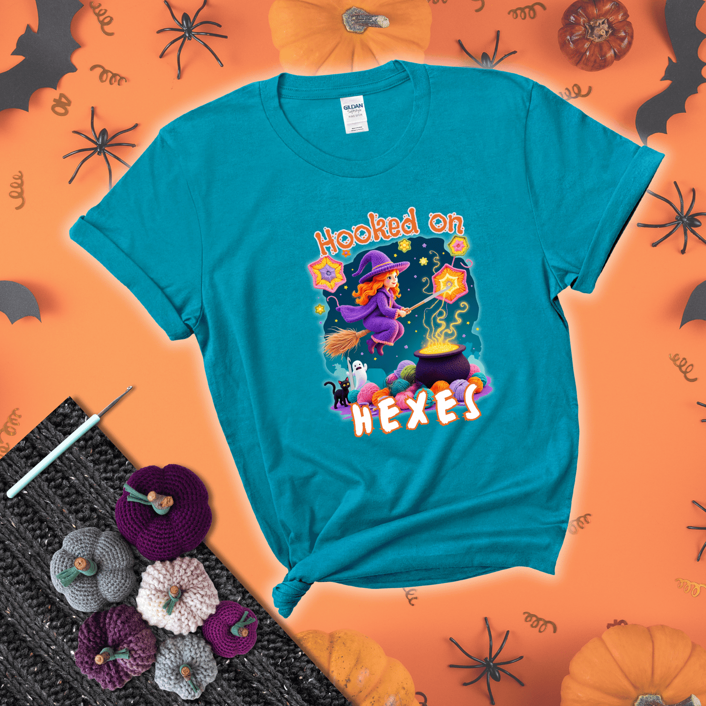 Witchy vibe with stars, hexagons, and a crochet hook. Text reads: “Hooked on Hexes.” A mystical and moody tee for fans of spooky stitches showing a blue-green tee with a festive Halloween background