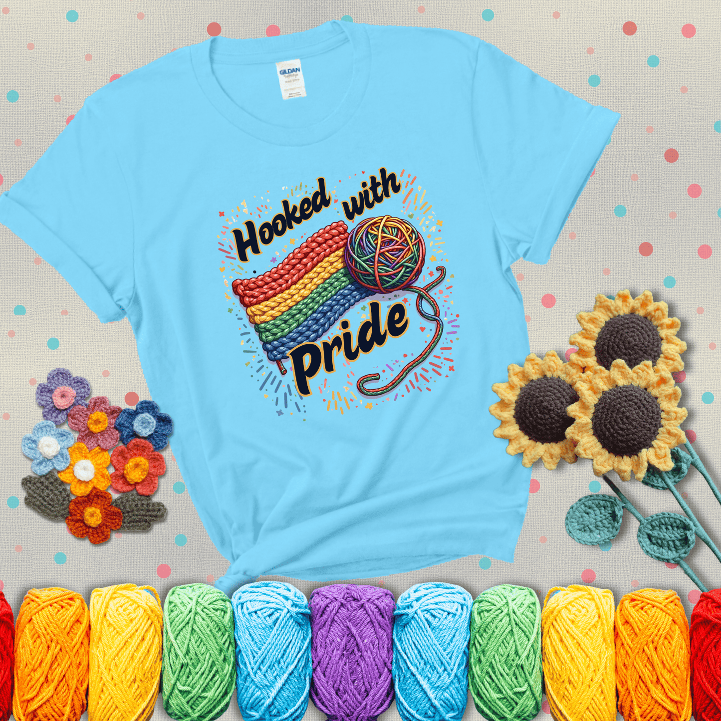 A rainbow-striped crochet swatch paired with a multicolor yarn ball and bold “Hooked With Pride” lettering. Confetti surrounds the vibrant scene showing a light blue tee with grey sunflower background