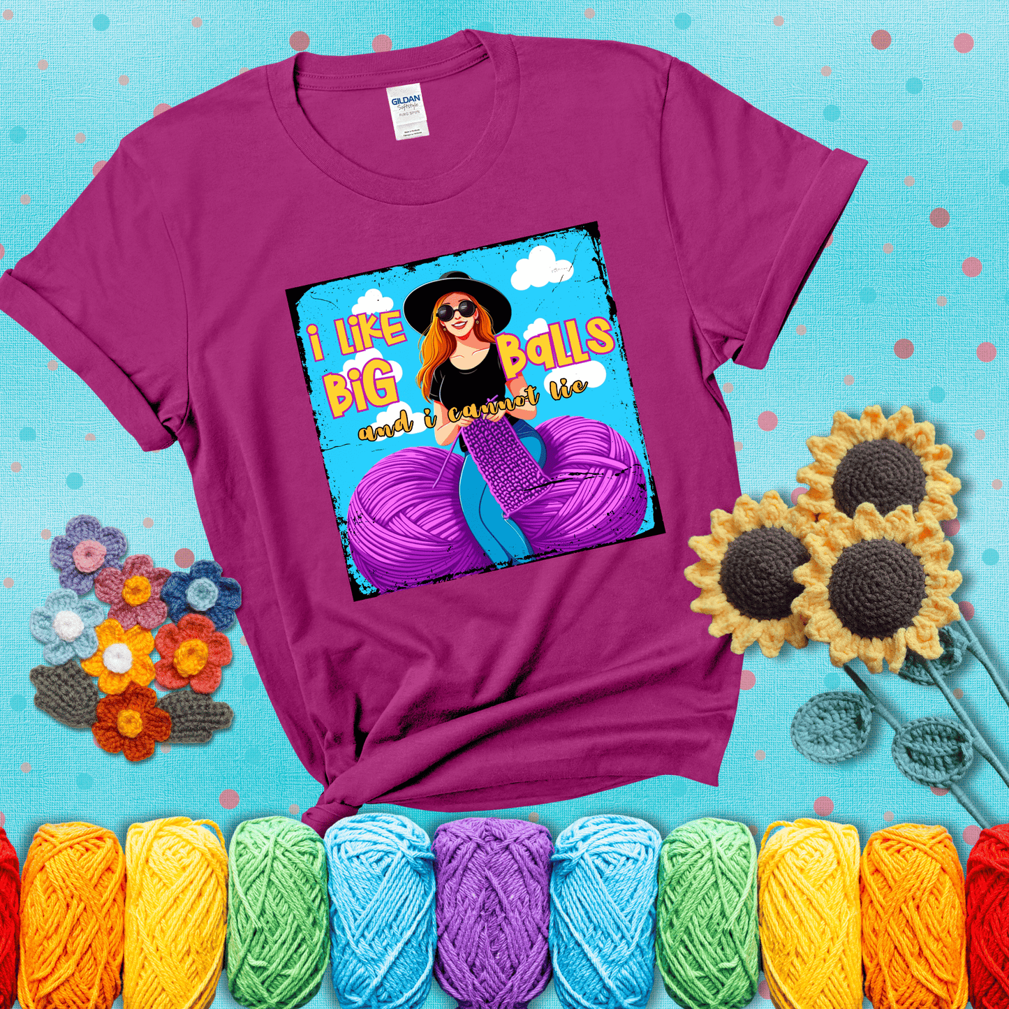 Bold cartoon image of a woman crocheting between two giant purple yarn balls with the phrase “I like big balls and I cannot lie” in retro fonts showing a magenta tee with a teal sunflower background