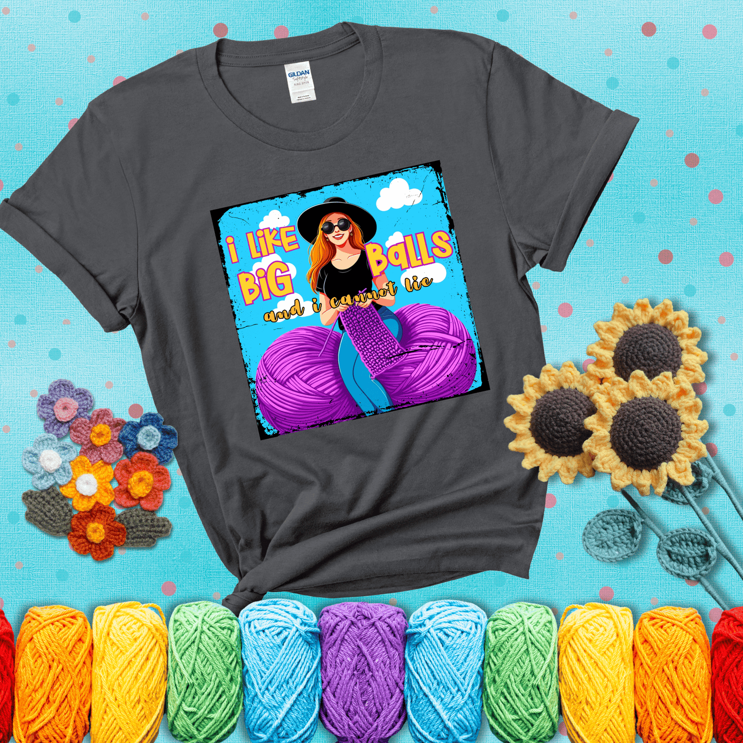Bold cartoon image of a woman crocheting between two giant purple yarn balls with the phrase “I like big balls and I cannot lie” in retro fonts showing a dark grey tee with a teal sunflower background