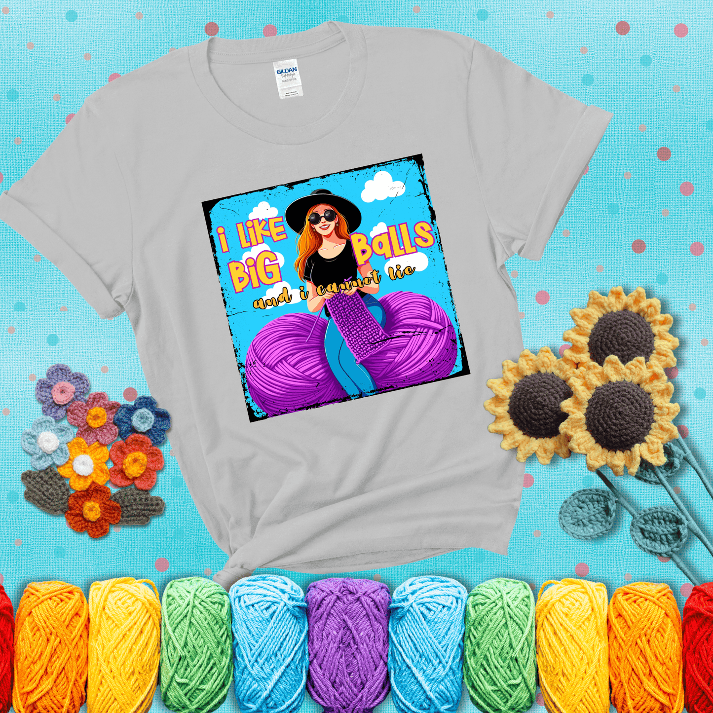 Bold cartoon image of a woman crocheting between two giant purple yarn balls with the phrase “I like big balls and I cannot lie” in retro fonts showing a light grey tee with a teal sunflower background