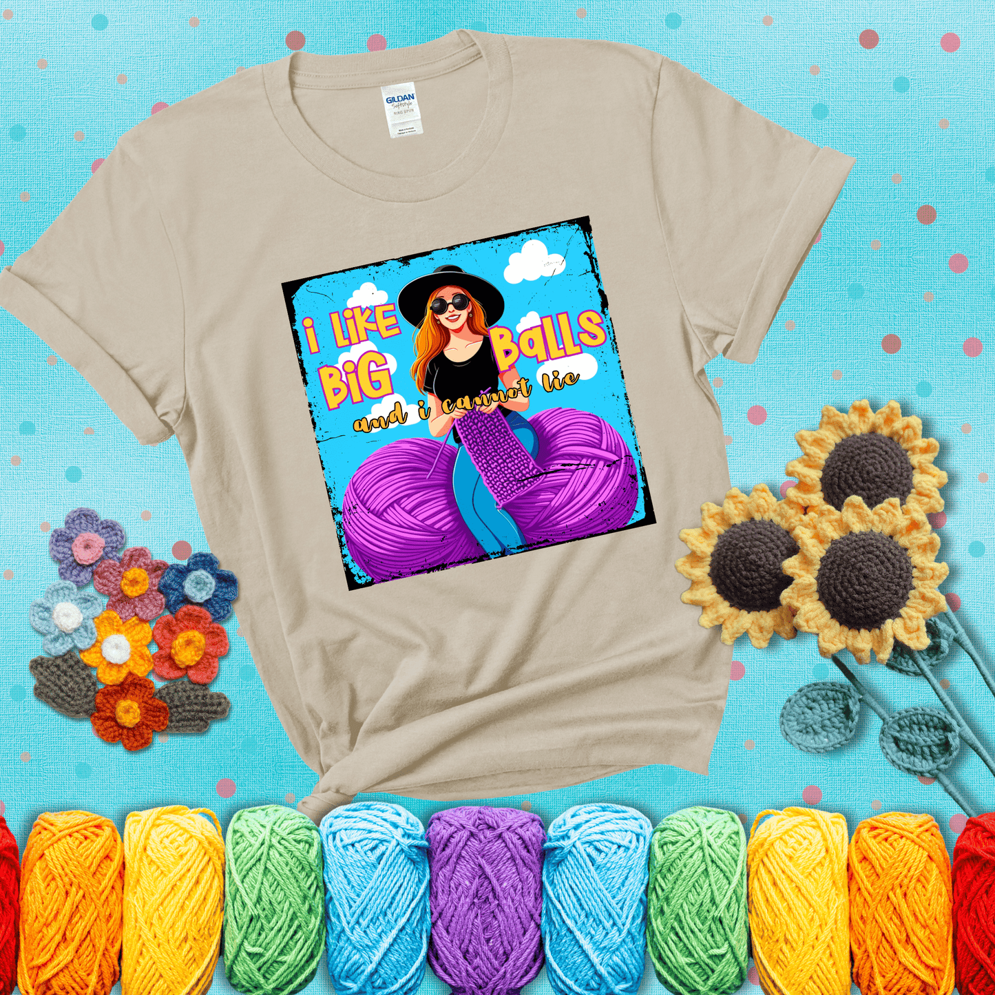 Bold cartoon image of a woman crocheting between two giant purple yarn balls with the phrase “I like big balls and I cannot lie” in retro fonts showing a beige tee with a teal sunflower background