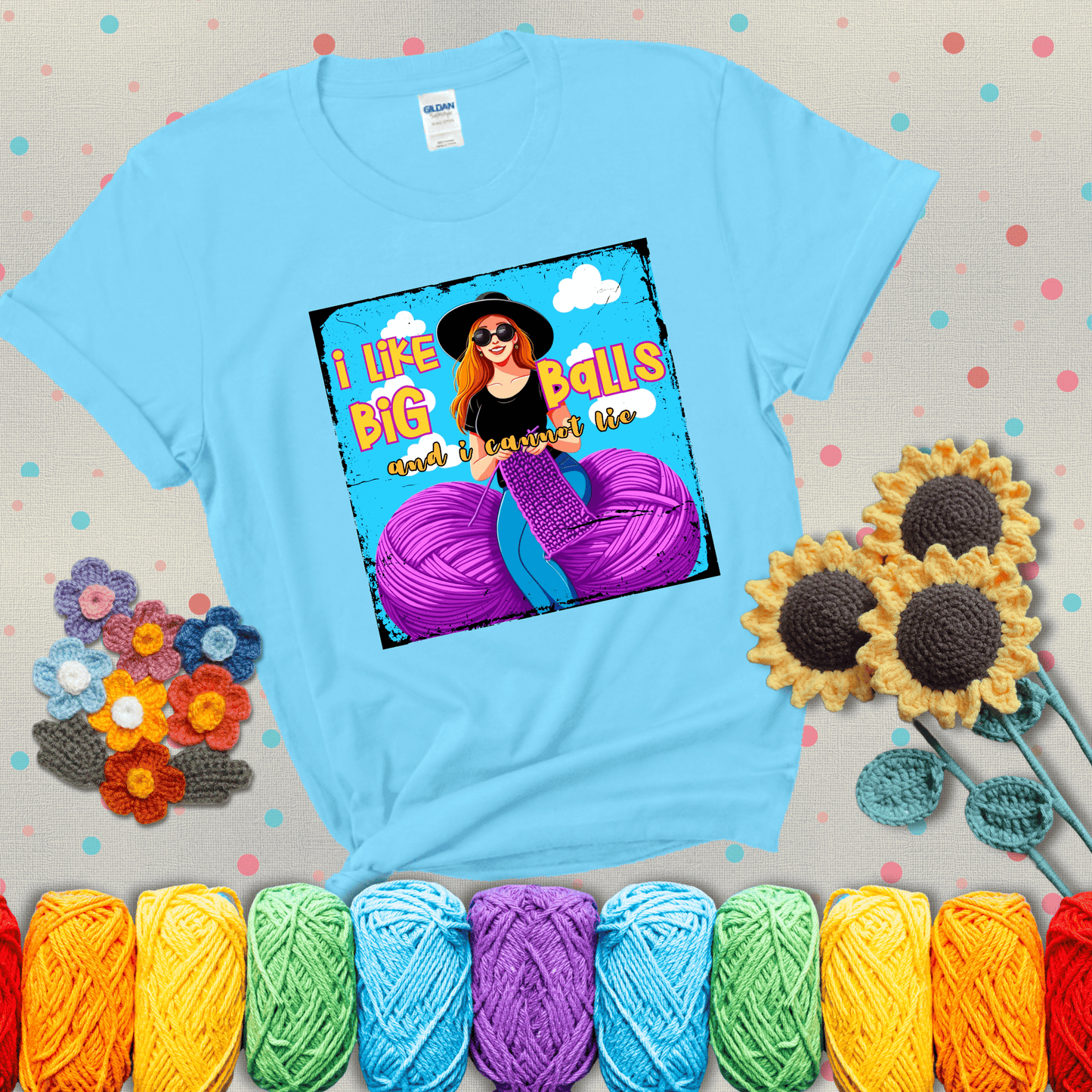 Bold cartoon image of a woman crocheting between two giant purple yarn balls with the phrase “I like big balls and I cannot lie” in retro fonts showing a light blue tee with grey sunflower background