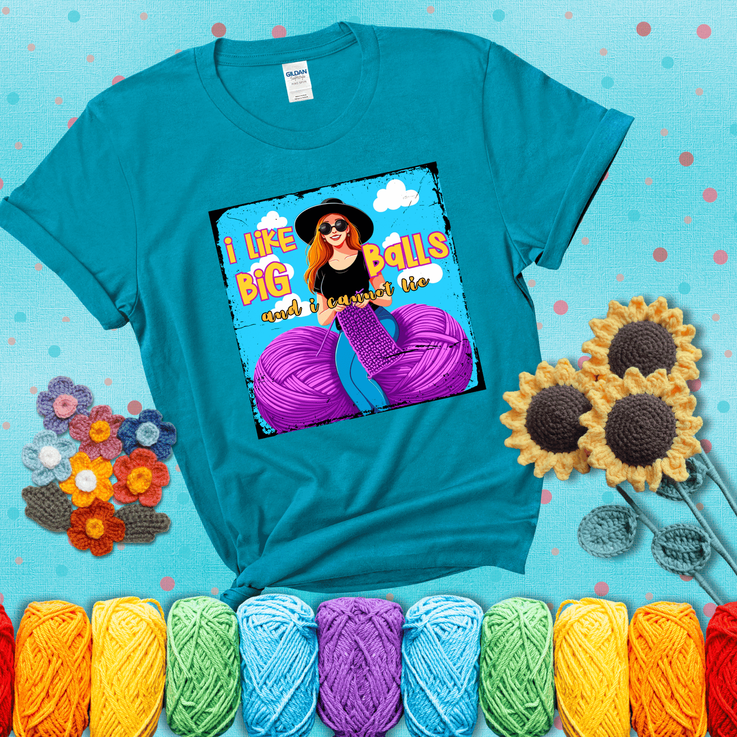 Bold cartoon image of a woman crocheting between two giant purple yarn balls with the phrase “I like big balls and I cannot lie” in retro fonts showing a blue-green tee with a teal sunflower background