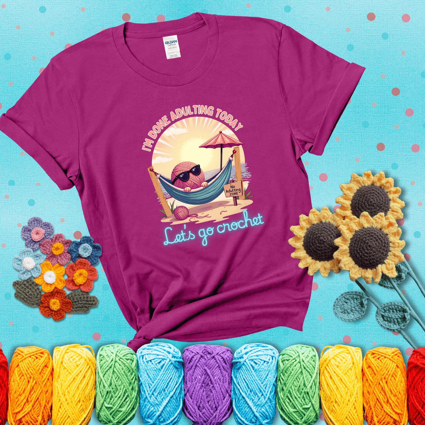 A chill yarn ball wearing sunglasses lounges in a hammock on a beach under the sun with the phrase “I’m done adulting today – Let’s go crochet,” showing a magenta tee with a teal sunflower background