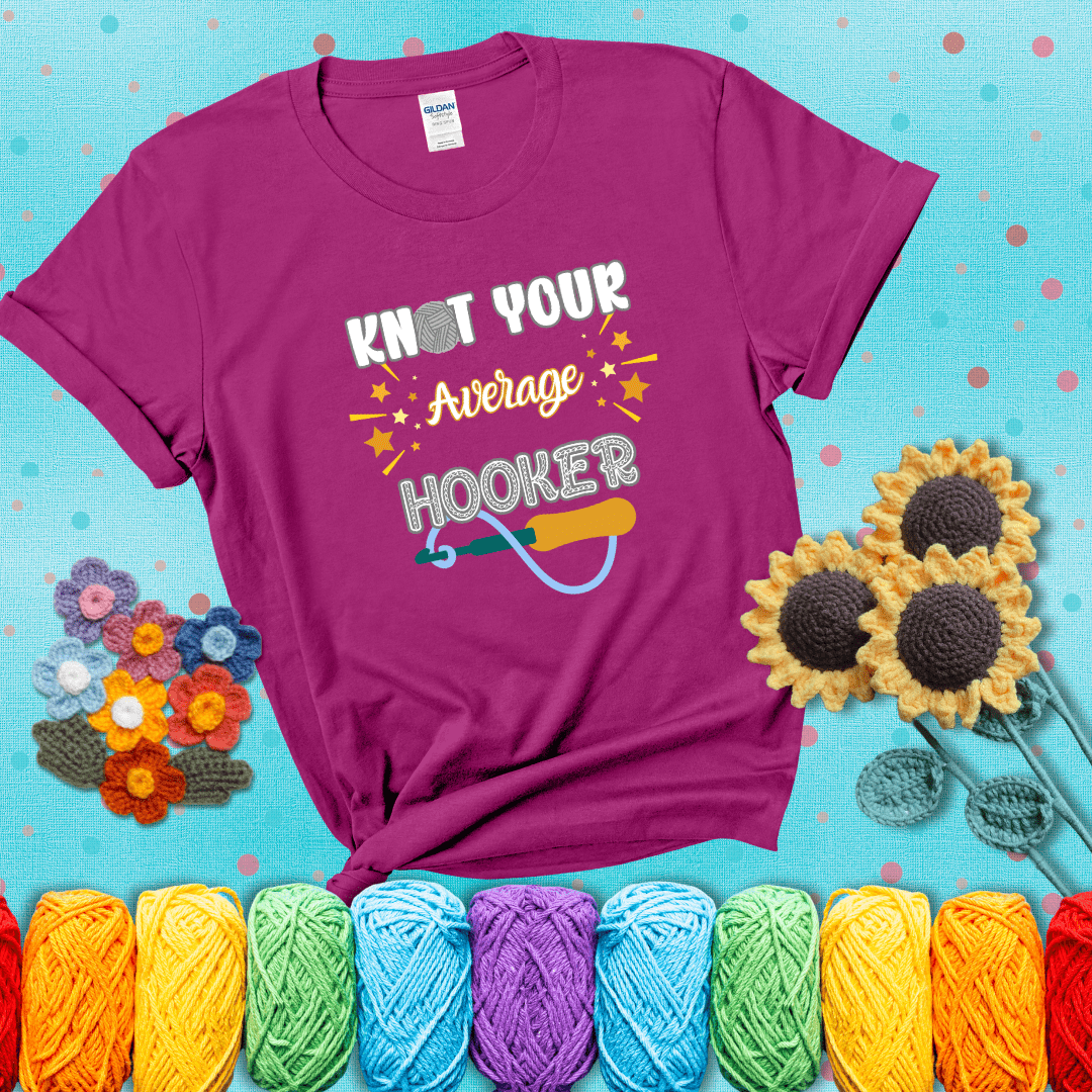 Bold lettering with a yarn hook graphic. Text reads: “I'm Not Your Average Hooker.” A cheeky crochet tee for confident crafters who own their skills, showing a magenta tee with a teal sunflower background
