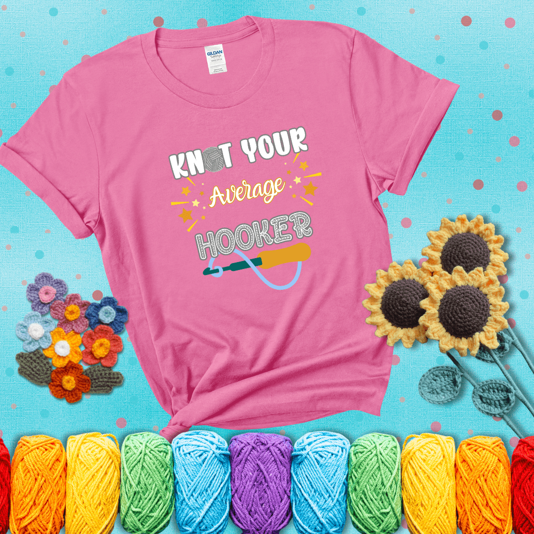 Bold lettering with a yarn hook graphic. Text reads: “I'm Not Your Average Hooker.” A cheeky crochet tee for confident crafters who own their skills, showing a pink tee with a teal sunflower background
